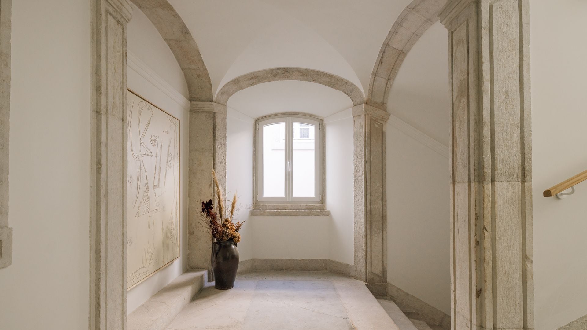 A corridor with a window, a vase with dried plants, and a door on the right.
