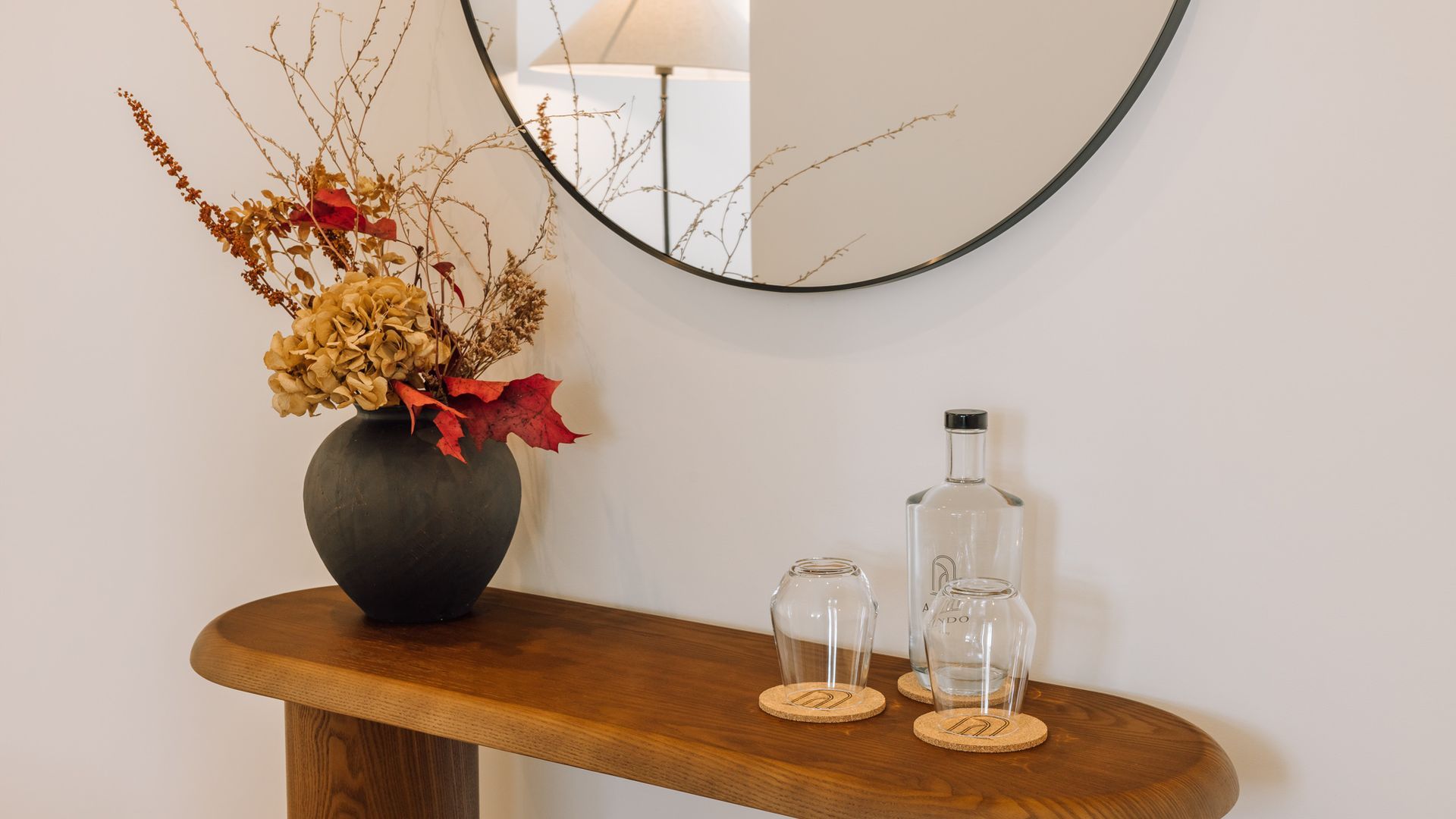 Een houten bijzettafeltje met een vaas gedroogde bloemen en twee heldere glazen flessen.