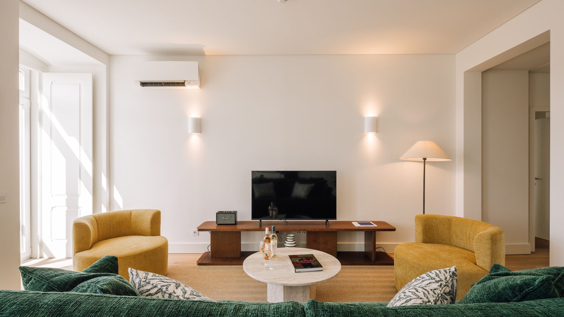 A modern living room with a flat-screen TV, two yellow armchairs, a round coffee table, and a sofa with green cushions.