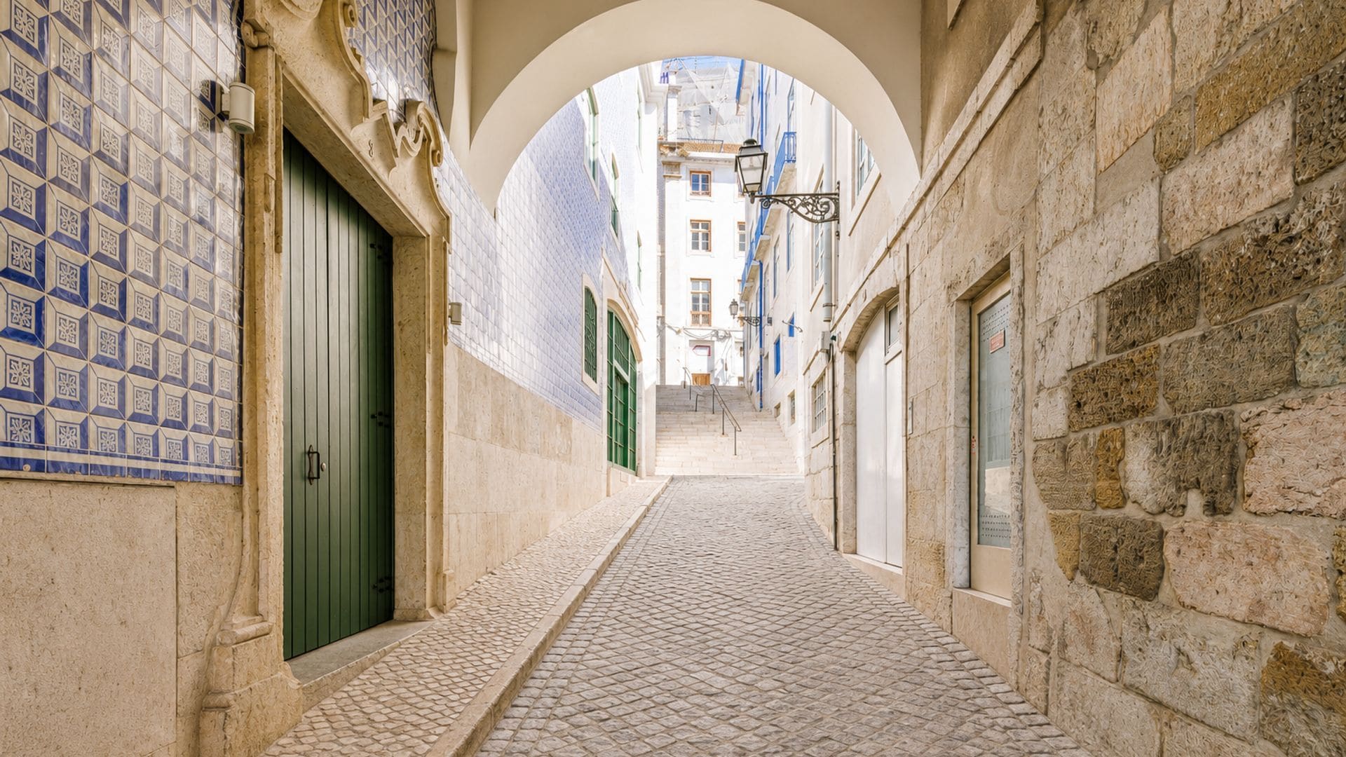 A narrow cobblestone alleyway with a tiled wall on the left and a stone wall on the right, leading to a building with a green door.