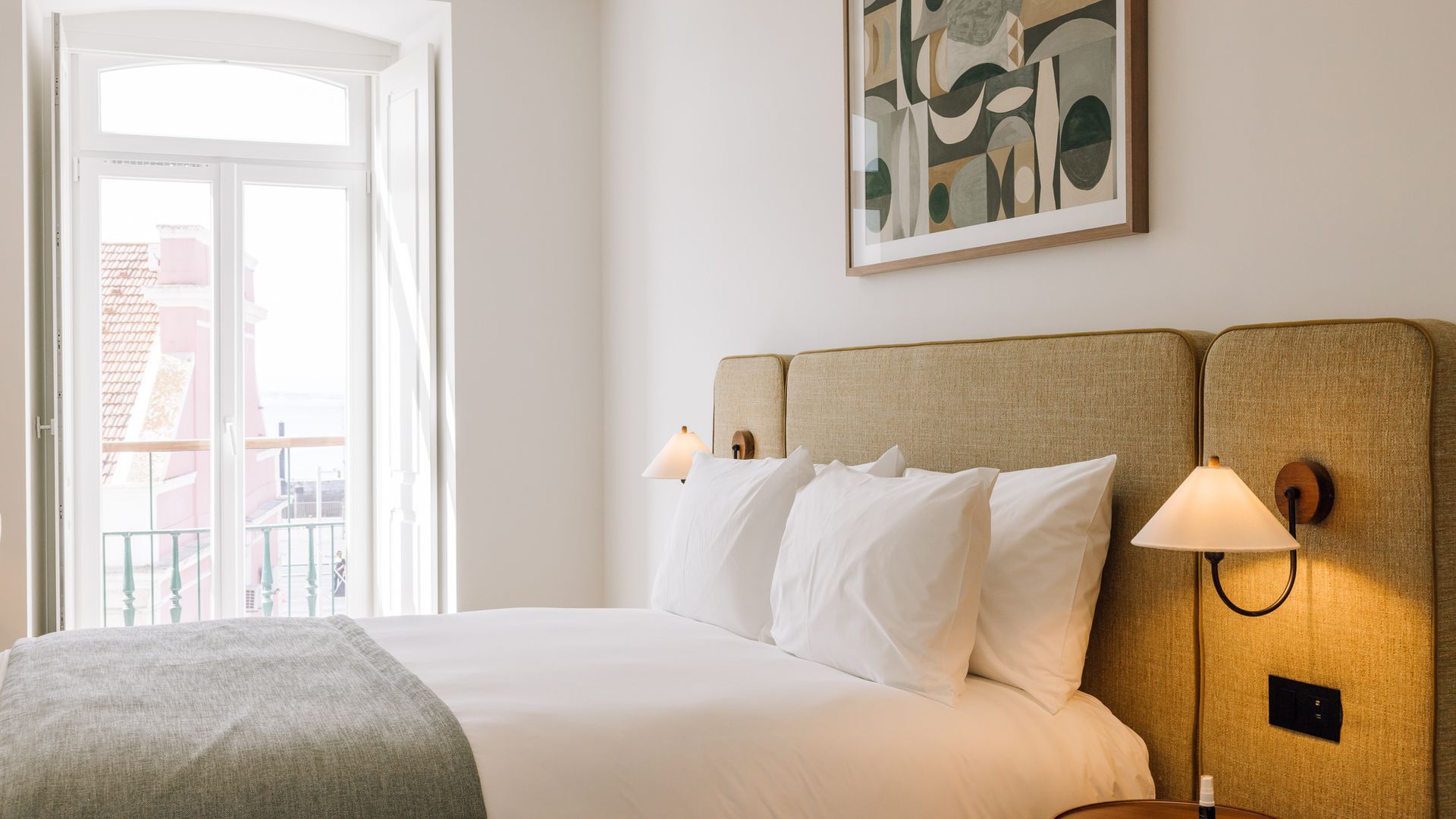 Une chambre d’hôtel avec un lit, deux lampes de chevet, une fenêtre donnant sur un paysage et un tableau au mur.