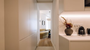 A narrow hallway leading to a dining area with a sofa and table, a vase with dried flowers, and a coffee maker on the counter. - Ando Living - Tejo Palace - 202 (Lisboa)
