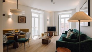 A modern living room with a sofa, a dining table set for two, a television, and a lamp. - Ando Living - Tejo Palace - 202 (Lisboa)