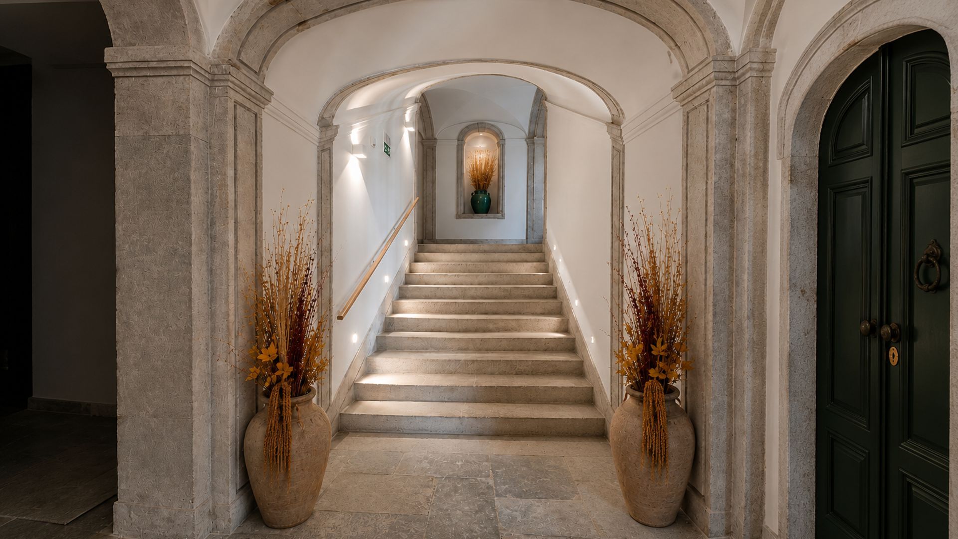 A grand staircase with a handrail, flanked by two large vases with dried plants, leading to a dark green door.