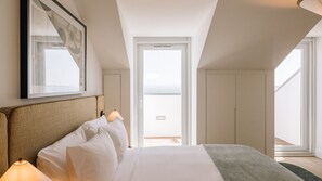 A hotel room with a bed, a nightstand, a lamp, a framed picture, and a view of the outside. - Ando Living - Tejo Palace - 301 (Lisboa)