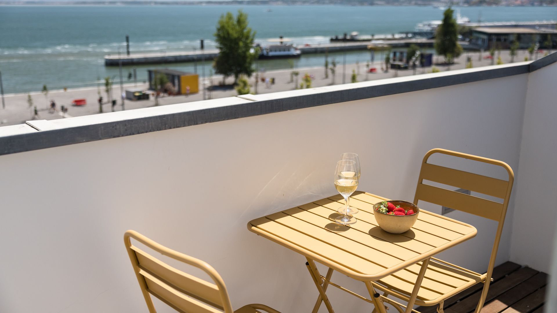 A balcony with a table and chairs, overlooking a waterfront with a promenade and buildings.