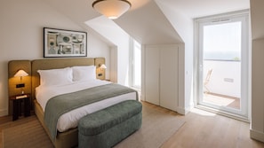 A hotel room with a bed, bedside lamps, a green throw, a patterned rug, a large window, and a balcony with a chair. - Ando Living - Tejo Palace - 301 (Lisboa)