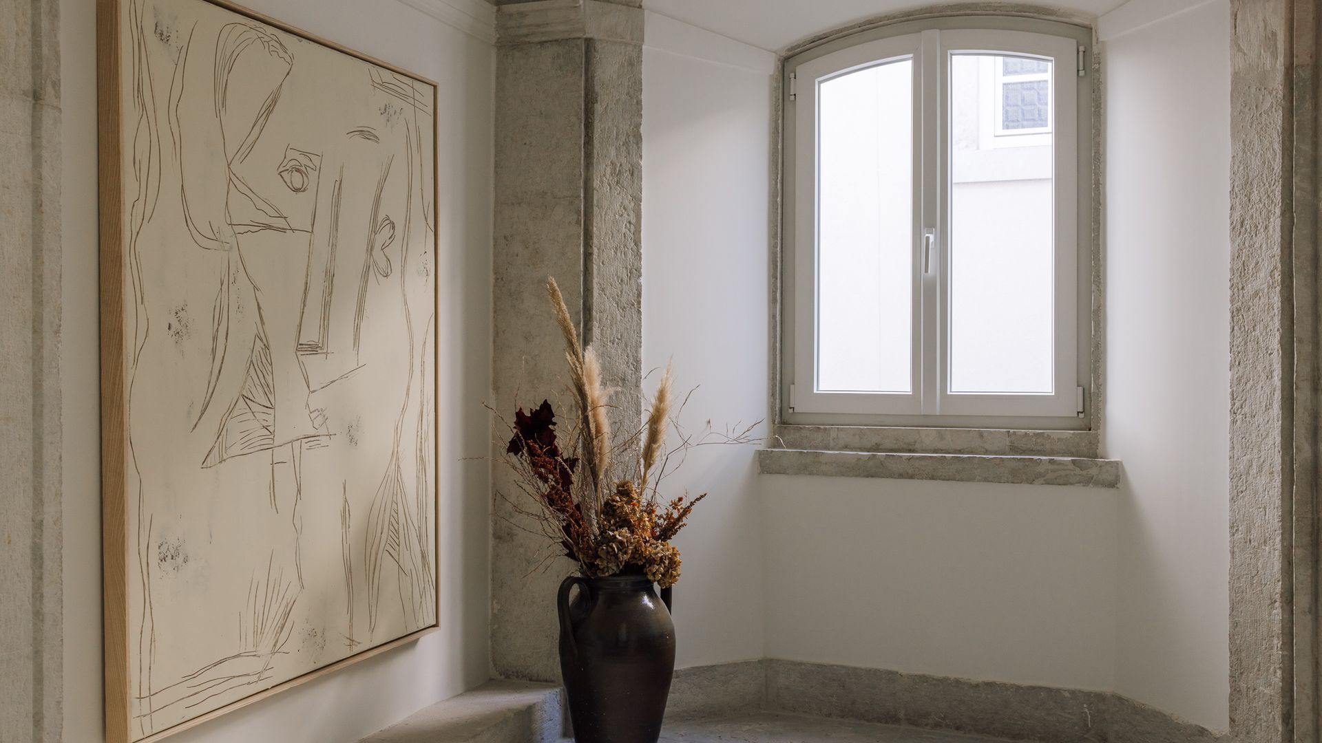 A room with a large abstract painting, a vase with dried flowers, and a window with a view.