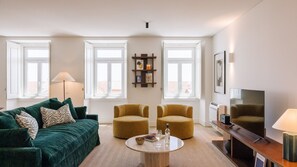 A living room with a green sofa, two armchairs, a coffee table, and a TV. There are two windows with white frames and a wall-mounted shelf with books and a mug. - Ando Living - Tejo Palace - 302 (Lisboa)