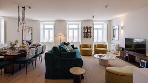 A modern living room with a sofa, armchairs, a coffee table, and a TV. There are dining tables with chairs and a window with a view. - Ando Living - Tejo Palace - 302 (Lisboa)
