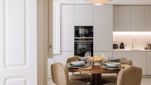A modern kitchen with white cabinetry, a built-in oven, and a dining area with wicker chairs and a round table set for four. - Ando Living - Tejo Palace - 303 (Lisboa)