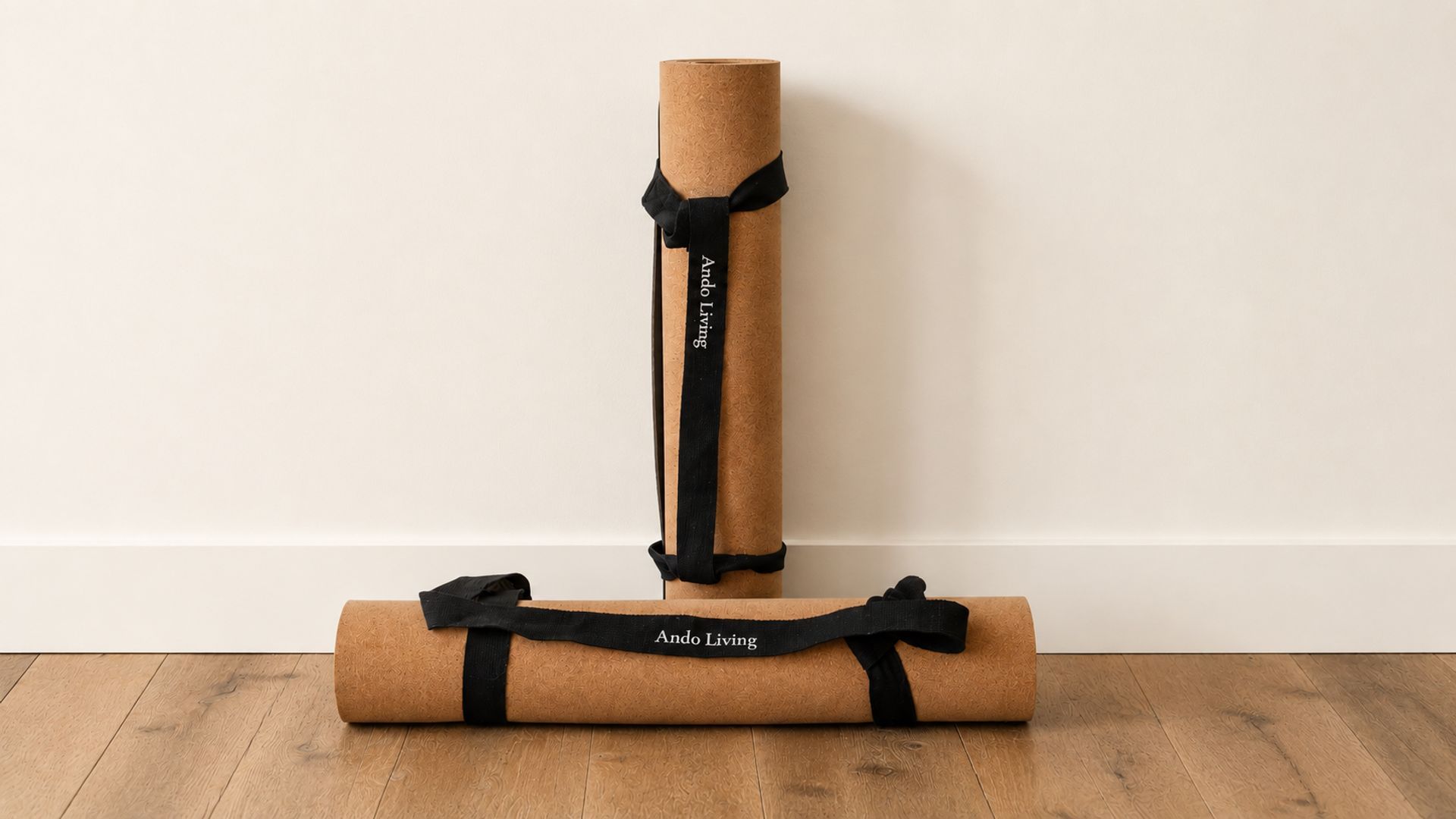 Twee yogamatten met 'Ando Living'-logo, waarvan één opgerold en één plat, op een houten vloer voor een effen muur.