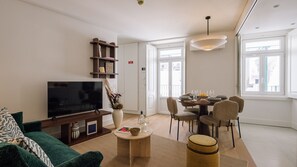 Uma sala de estar moderna com uma TV de tela plana, uma mesa de centro com um vaso, um espaço de refeições com cadeiras e uma janela grande.
