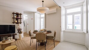 A modern living room with a flat-screen TV, a dining area with a round table and wicker chairs, and large windows providing natural light. - Ando Living - Tejo Palace - 303 (Lisboa)