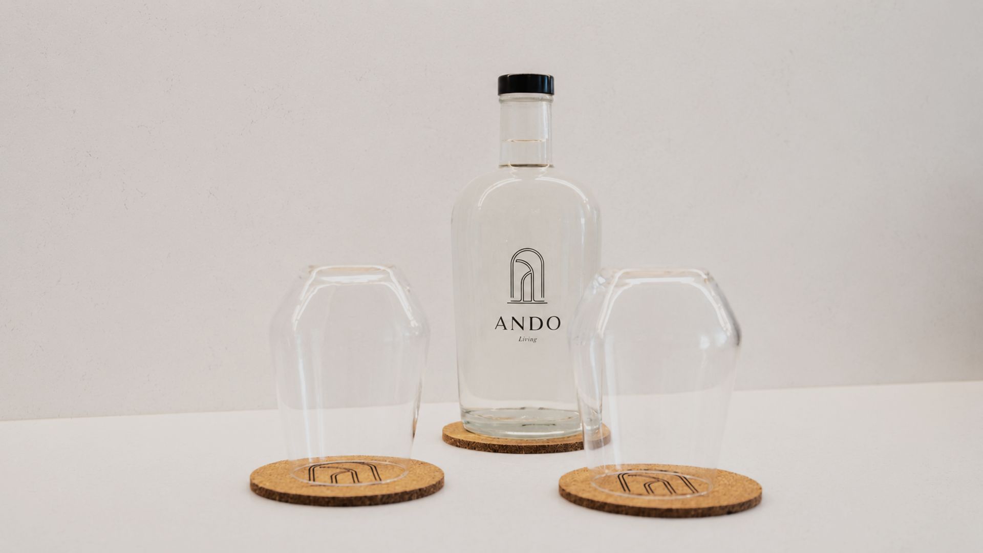 Een transparante fles met zwarte dop en een logo, twee transparante glazen en drie kurken onderzetters met een logo op een witte ondergrond.