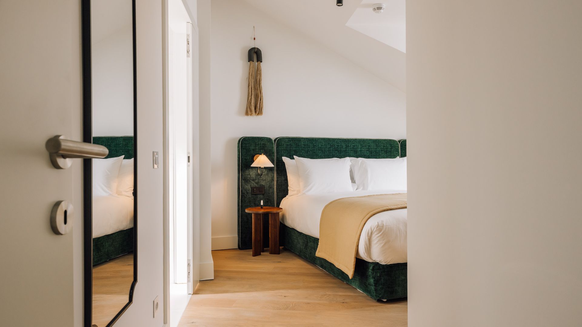 Hotellihuone, jossa on peilillä varustettu ovi, vihreällä päätyosalla varustettu sänky, yöpöytä lampun kanssa ja katosta roikkuva valaisin.