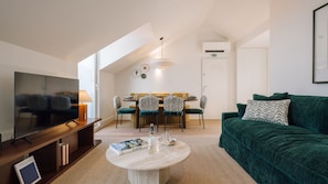 A modern living room with a flat-screen TV, a green sofa, a dining area with a yellow sofa and patterned chairs, and a large window providing natural light. - Ando Living - Tejo Palace - 400 (Lisboa)
