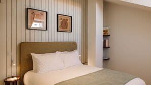 Quarto de hotel com cama, mesa de cabeceira, um quadro emoldurado e uma estante com livros.