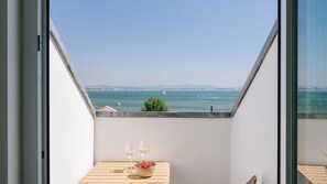 Varanda com mesa posta para dois, com vista para um corpo de água e um veleiro ao longe.
