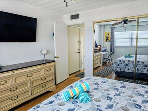 Un dormitori amb un llit, una petxera, un escriptori, una cadira, un televisor i una porta que dona a una altra habitació.