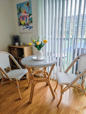 Una habitació amb una taula rodona, dues cadires de vimetes, un vas blanc amb flors grogues, una màquina de cafè i un quadre a la paret.