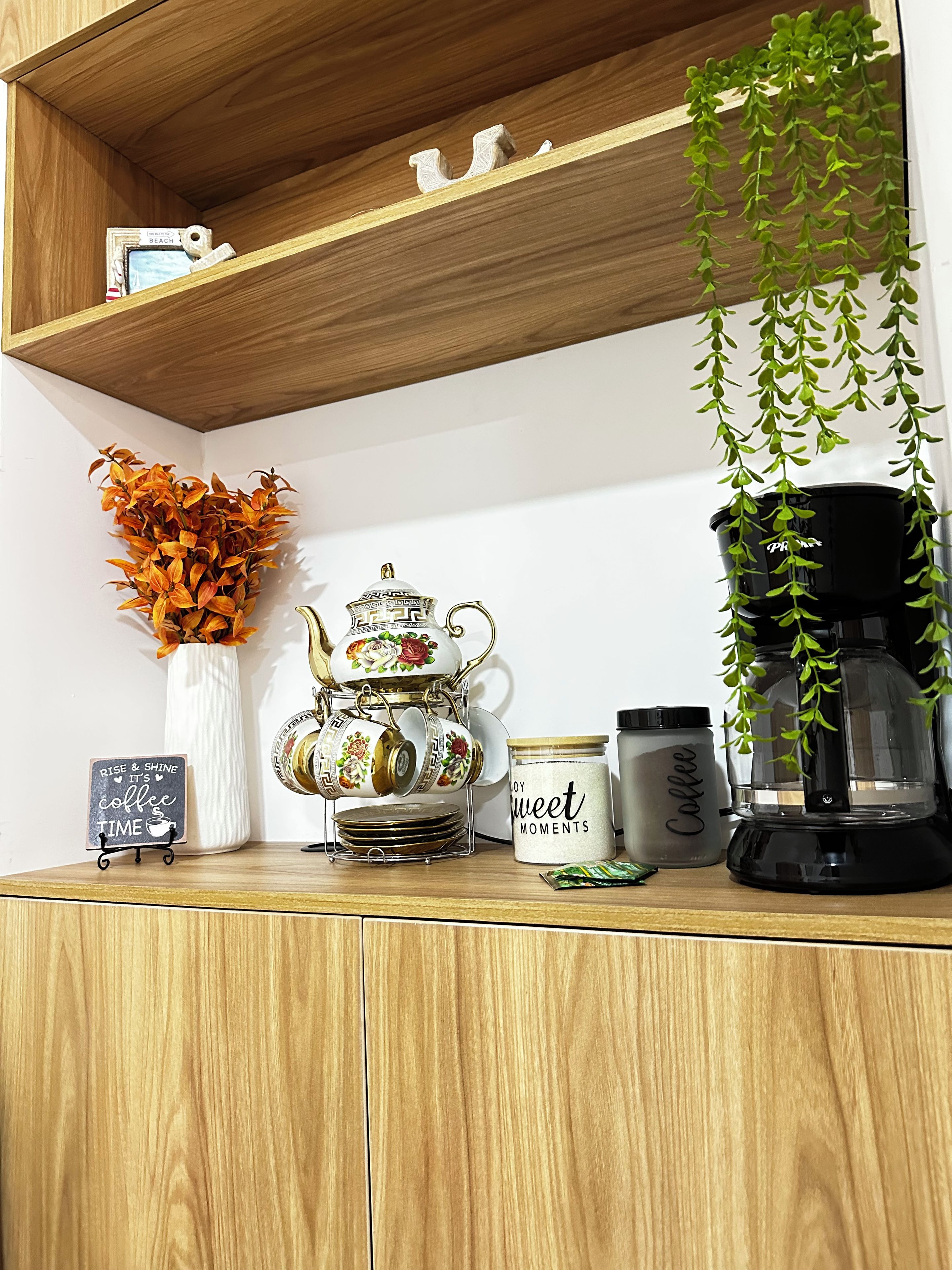 Een houten plank met een decoratieve theepot, een koffiezetapparaat, een potje met de tekst 'Koffie' en een bordje waarop staat 'Opstaan en Schijnen, het is koffietijd'.
