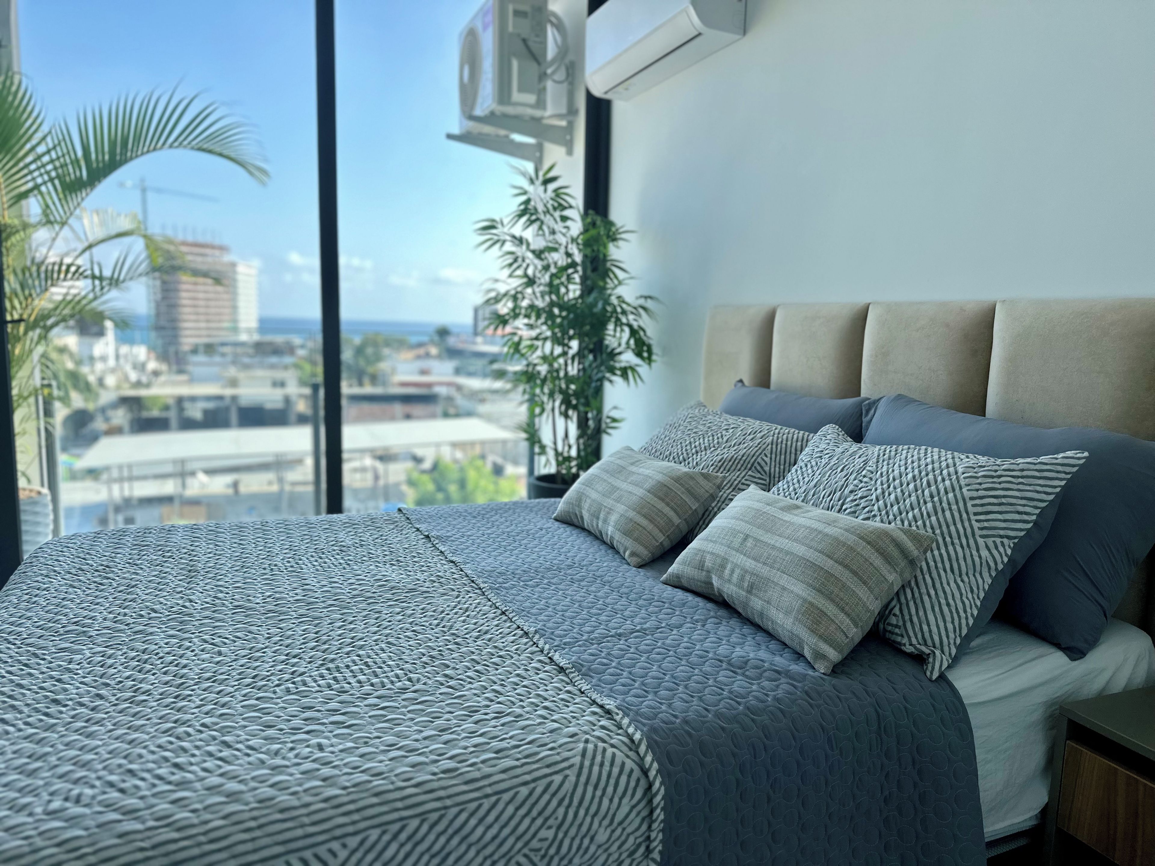 A bed with a textured bedspread and multiple pillows, a window with a view of buildings and palm trees, and an air conditioning unit mounted on the wall.