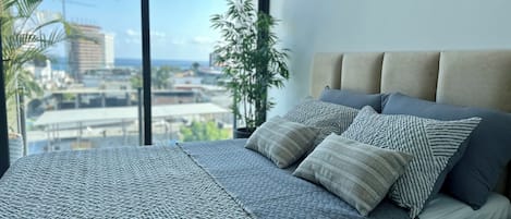 A bed with a textured bedspread and multiple pillows, a window with a view of buildings and palm trees, and an air conditioning unit mounted on the wall.