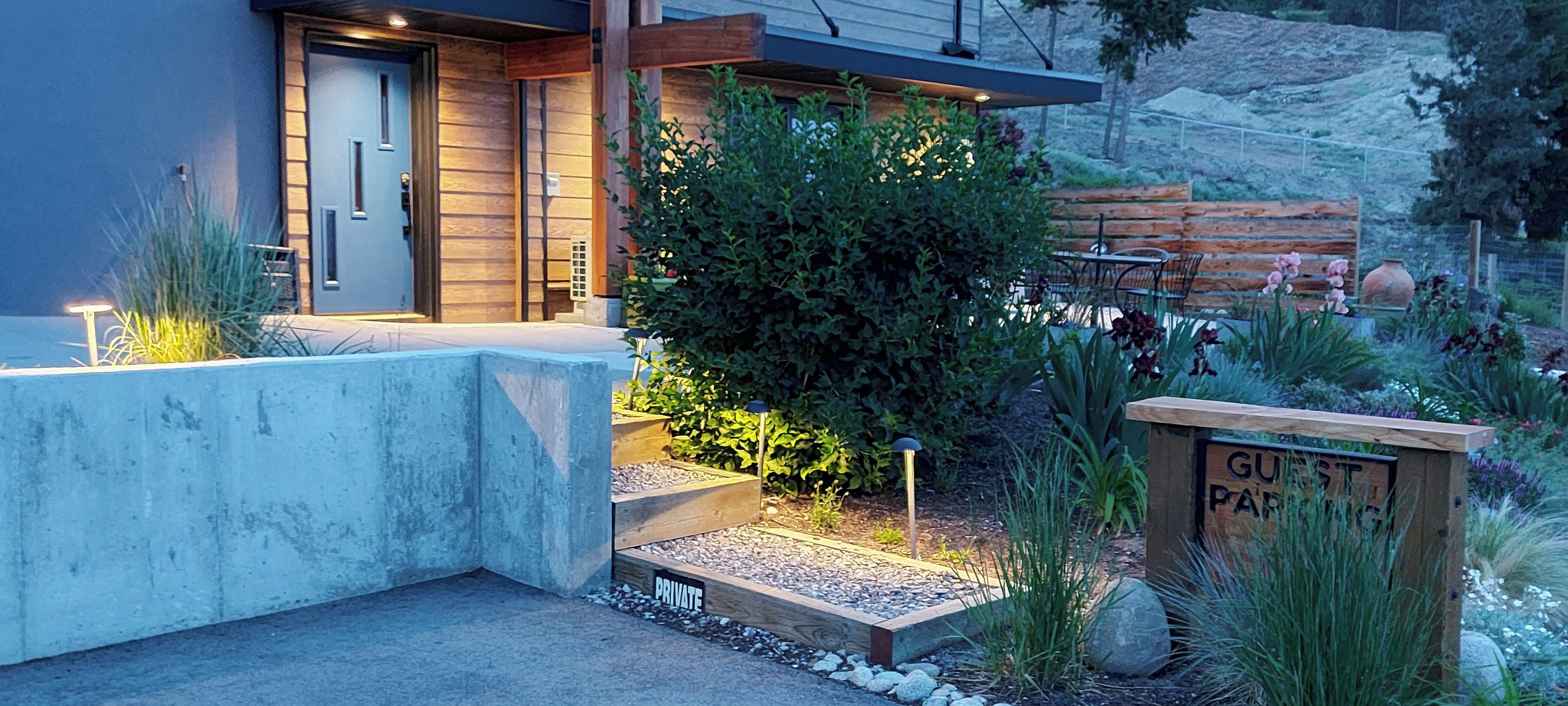 A private garden entrance with a concrete pathway leading to a wooden deck, surrounded by lush greenery and a sign indicating 'PRIVATE GUEST PARKING'.