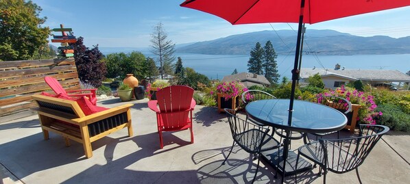An outdoor patio with a wooden bench, red chairs, a round table, and a red umbrella.