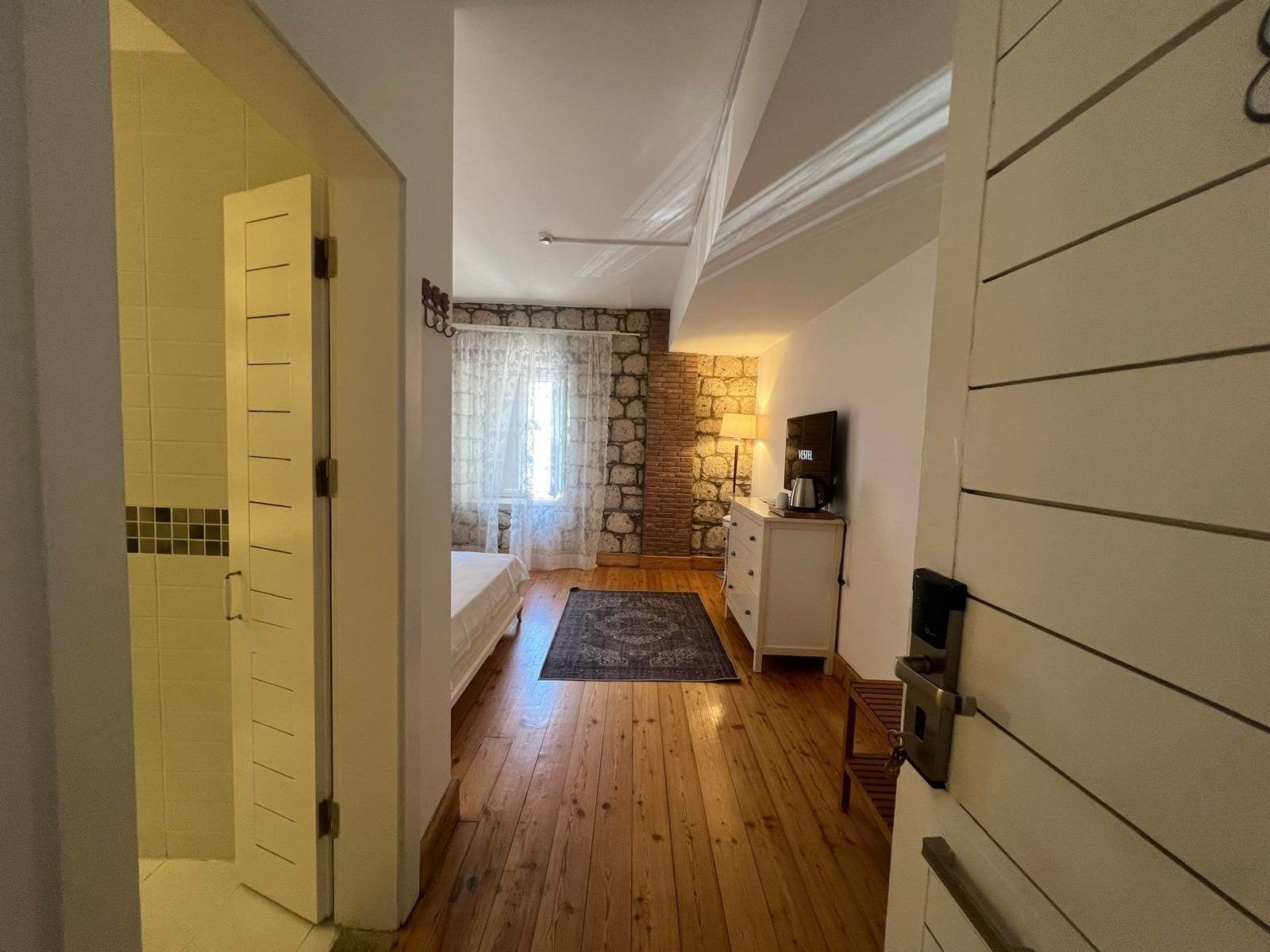 Un pasillo estrecho que conduce a un dormitorio con pared de piedra, una cama, un aparador y un televisor.