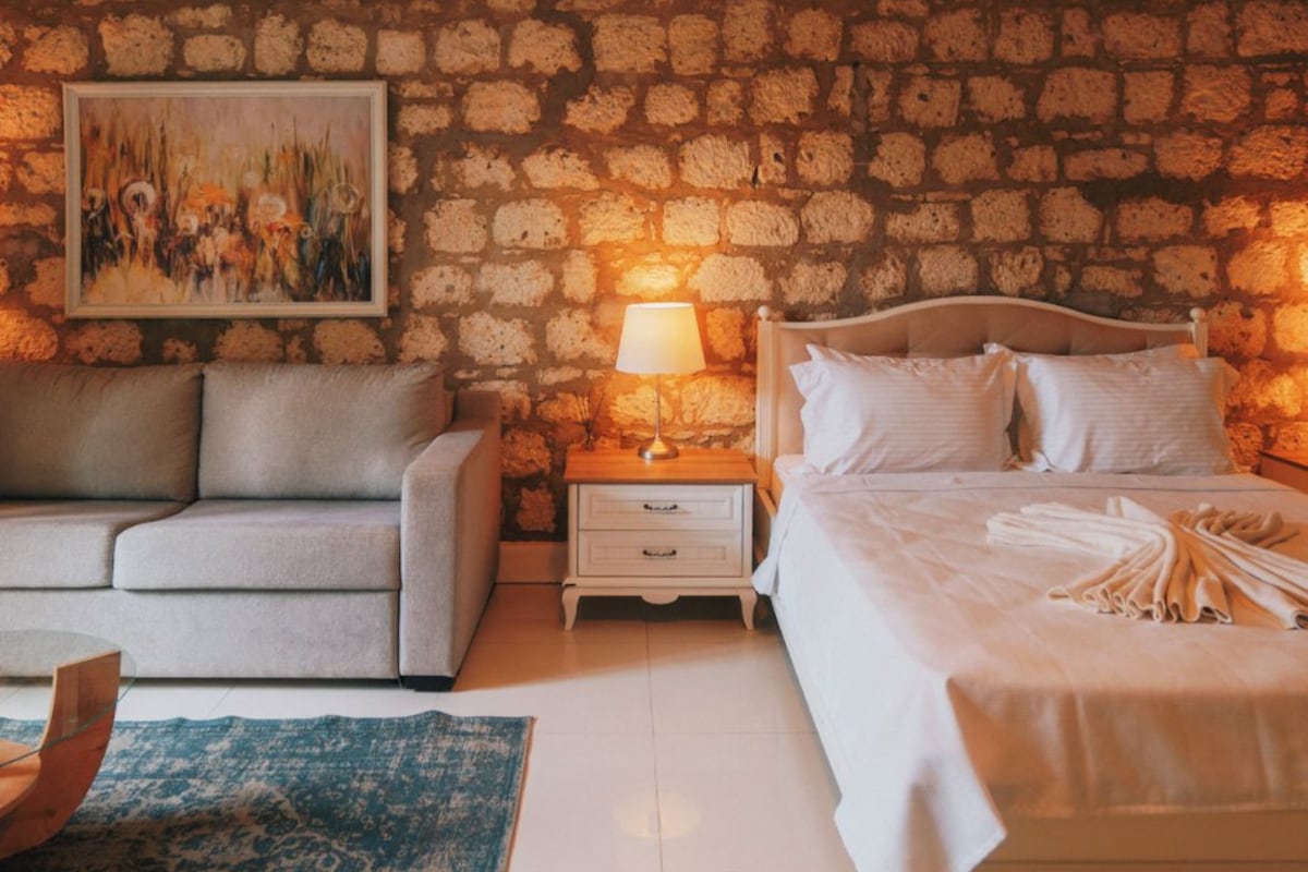Un dormitorio con pared de piedra, cama, mesita de noche, lámpara, sofá y un cuadro.