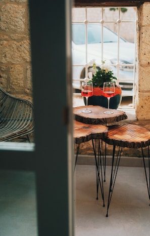 Una mesa de madera con dos vasos de líquido rojo, una silla de mimbre y una planta en maceta en un balcón con vista a un coche a través de una ventana.