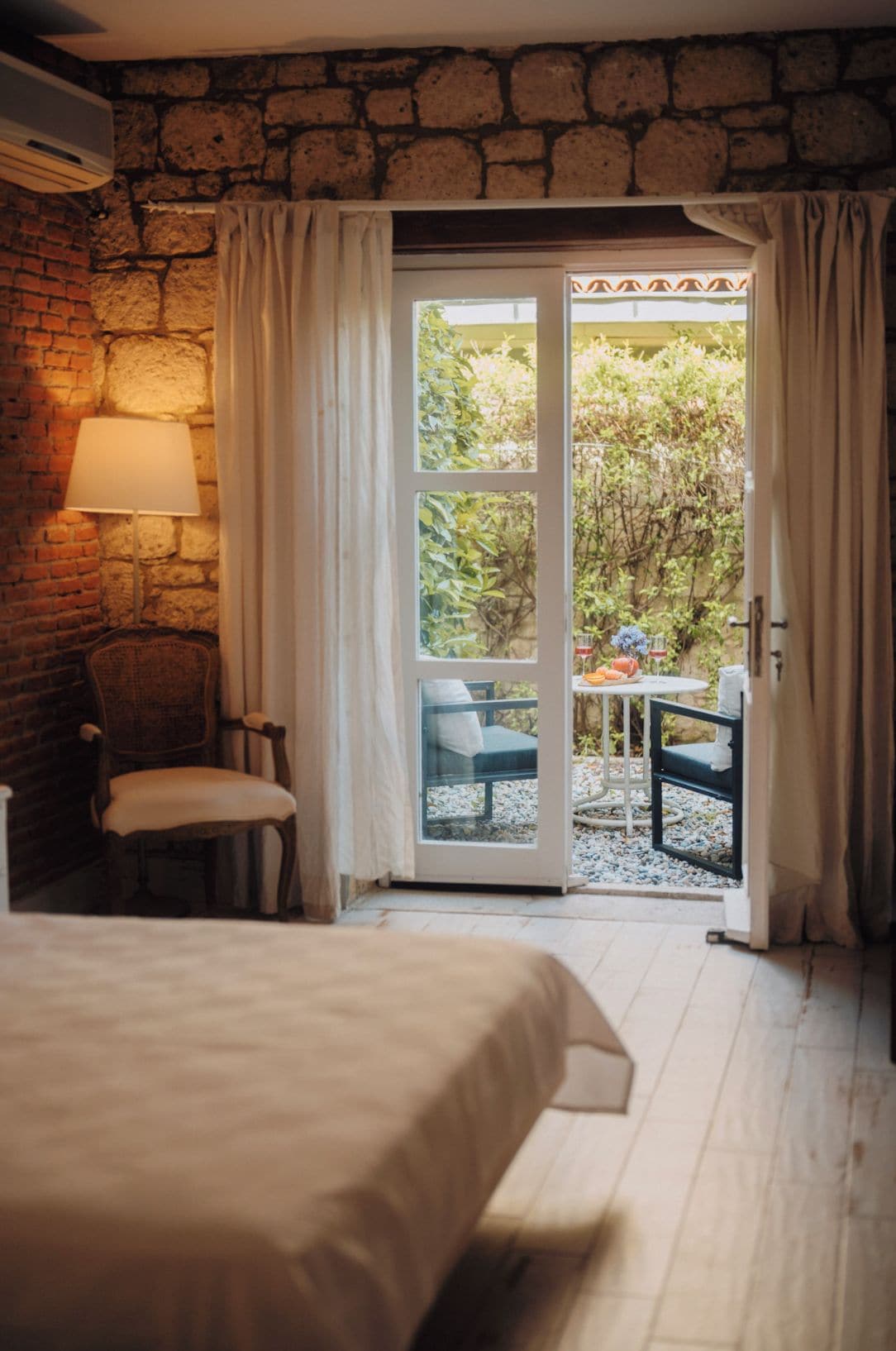 Una habitación con una cama, una silla, una mesa con otra silla afuera y una pared de piedra.