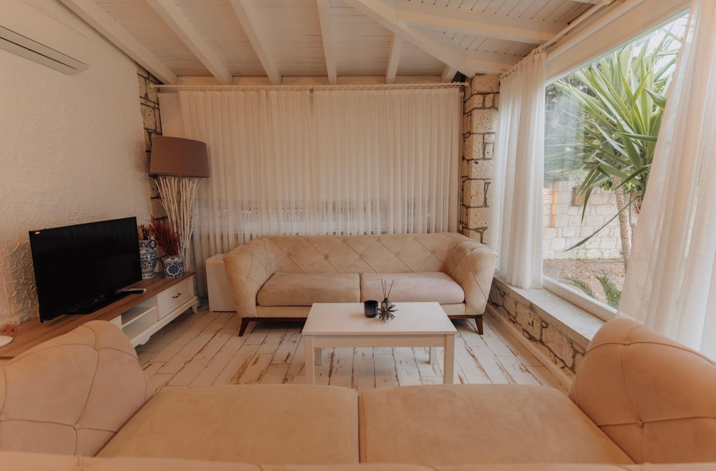 Una sala de estar con un sofá color beige, una mesa de centro, un televisor sobre un mueble de madera y una ventana con cortinas.