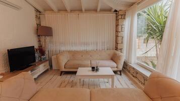 Una sala de estar con un sofá color beige, una mesa de centro, un televisor sobre un mueble de madera y una ventana con cortinas.