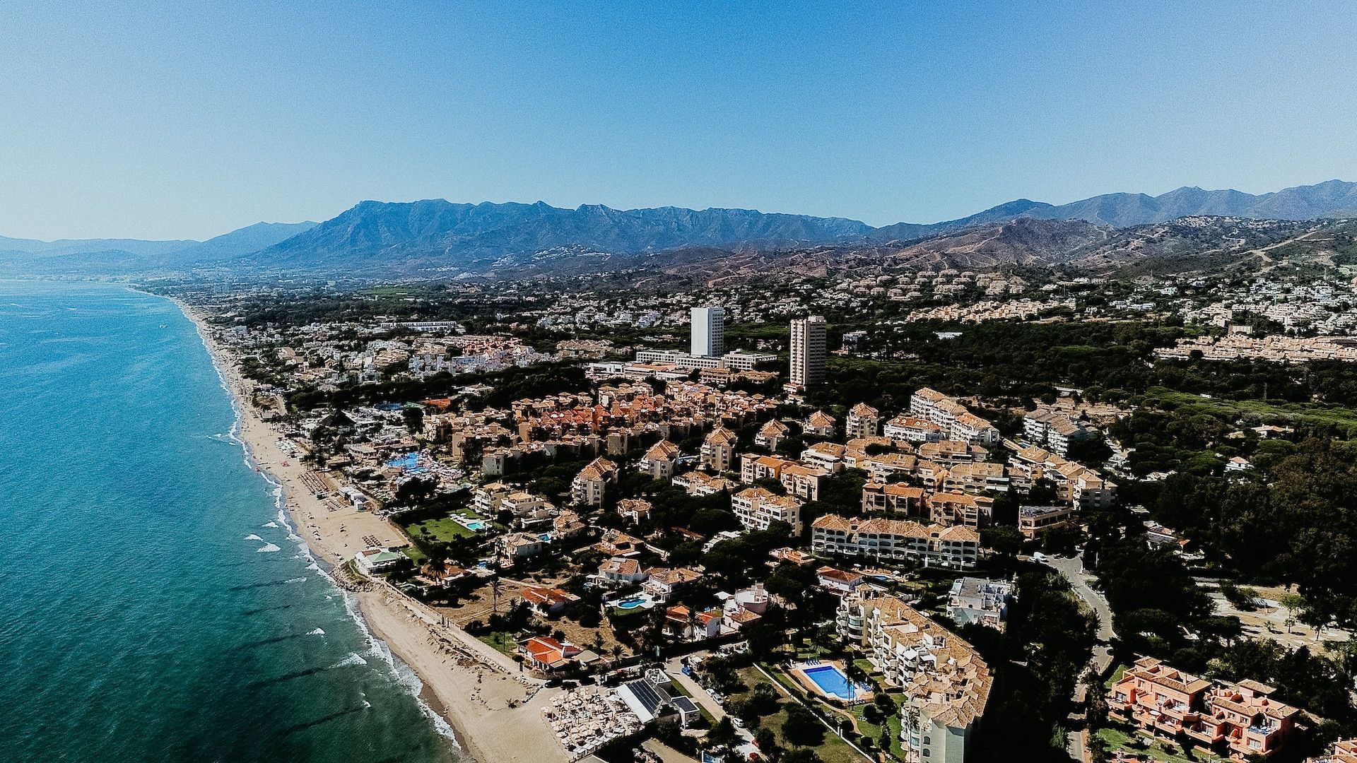 An aerial view of a coastal city with a beach, residential buildings, and mountains in the background.