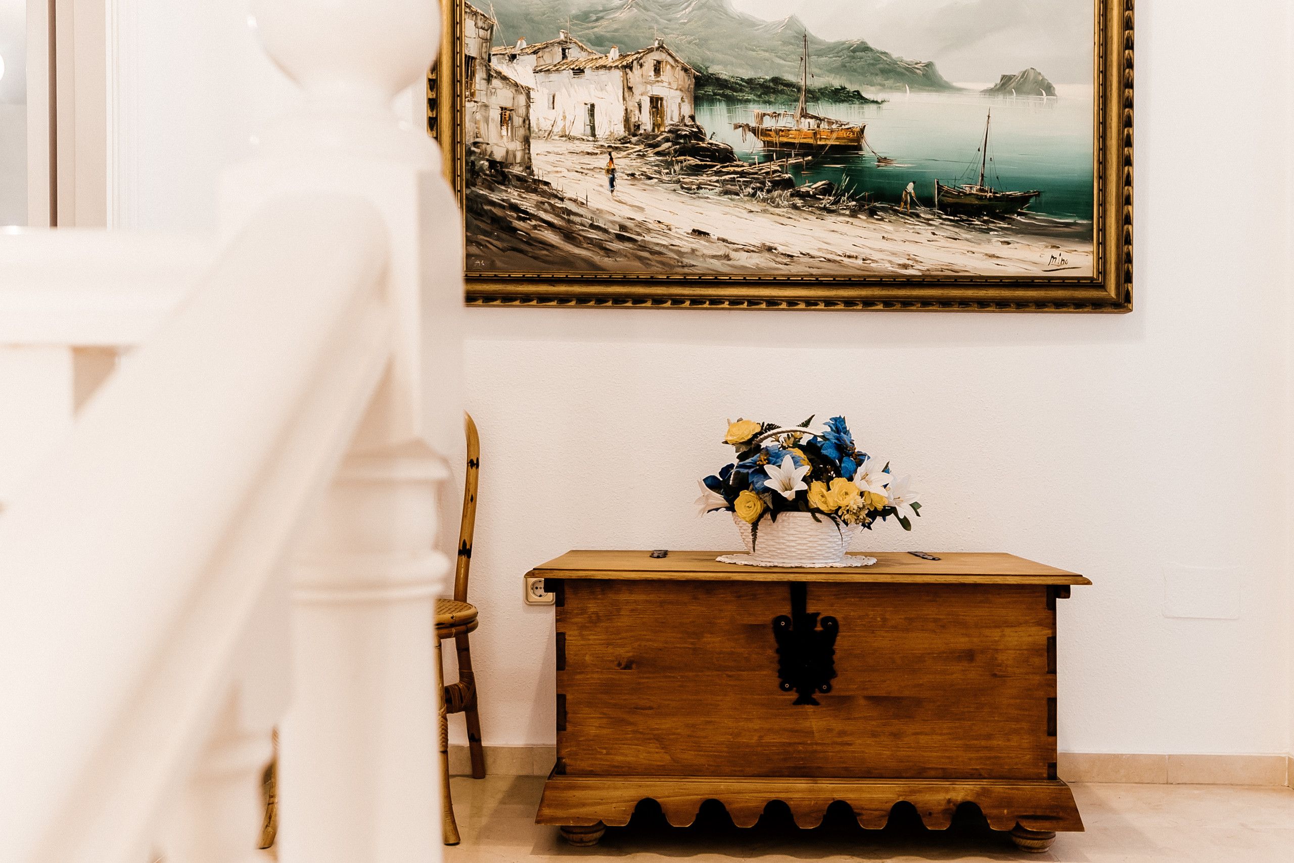 A room with a wooden dresser, a vase of flowers, a chair, and a painting on the wall.