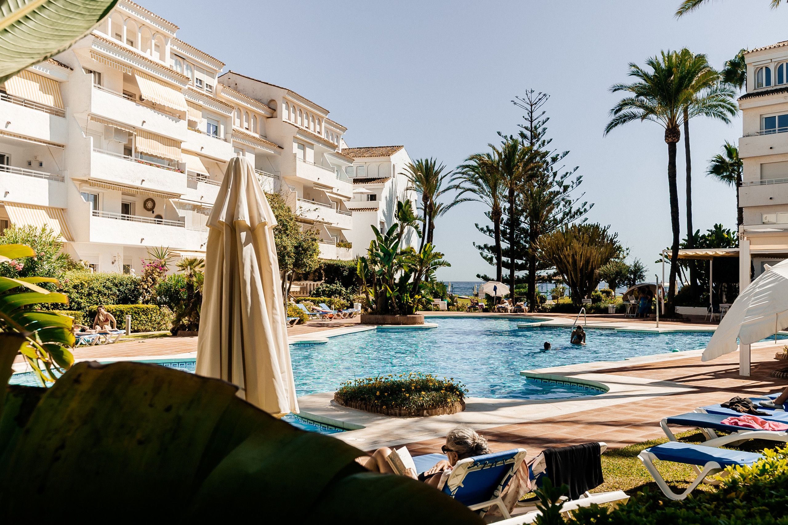 Un área de piscina con sillas de descanso, palmeras y un edificio con balcones.