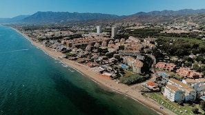 An aerial view of a coastal town with a beach, residential buildings, and mountains in the background. - 03 Beach Home by Abarzo (Marbella)