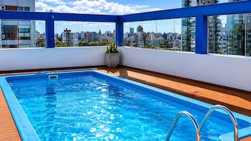 A rooftop swimming pool with a wooden deck, surrounded by a blue railing, and overlooking a cityscape with high-rise buildings.