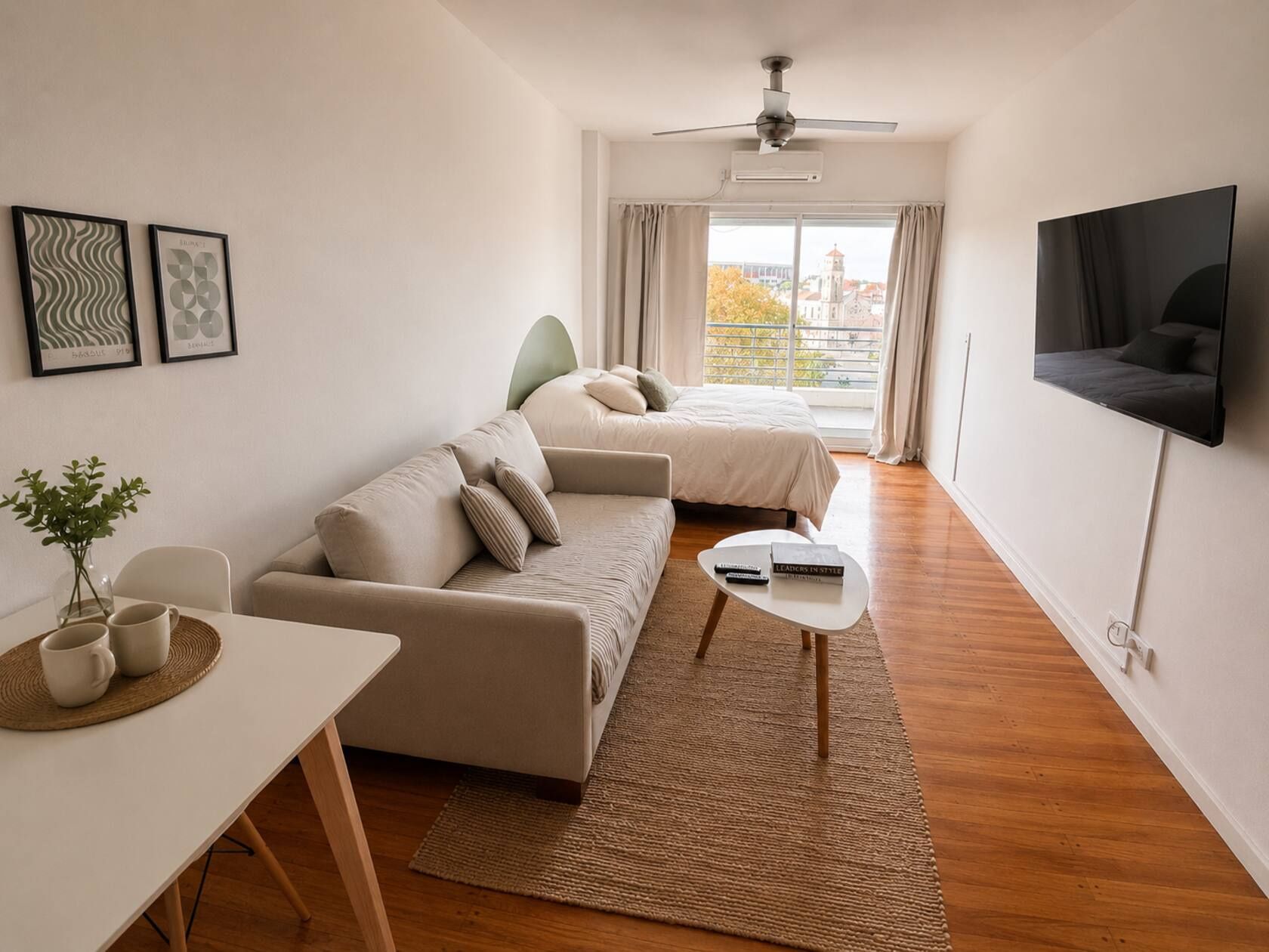 A modern hotel room with a sofa, a bed, a TV mounted on the wall, and a window with curtains.