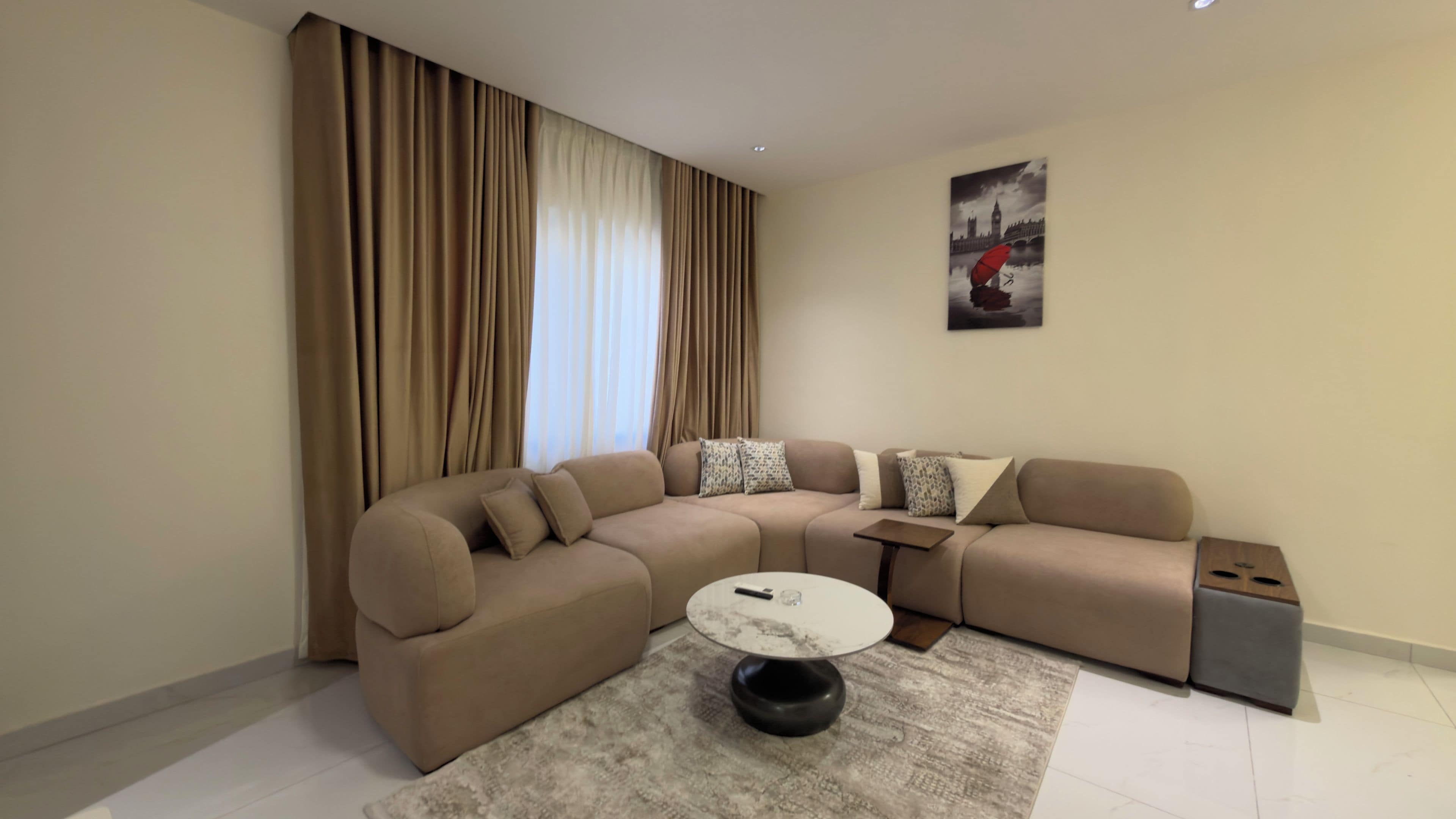 Een moderne woonkamer met een beige hoekbank, een ronde salontafel en een groot raam met gordijnen.