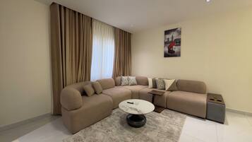 Een moderne woonkamer met een beige hoekbank, een ronde salontafel en een groot raam met gordijnen.