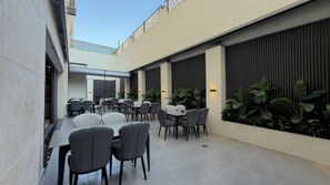A modern outdoor dining area with tables and chairs, surrounded by plants and a large window.