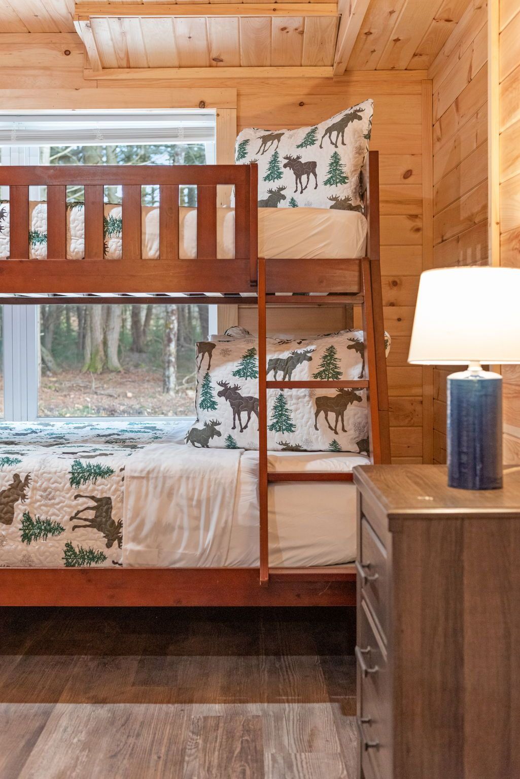 Uma cama beliche com roupa de cama com tema de floresta, uma luminária de cabeceira e vista para o exterior.