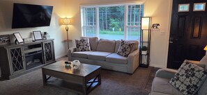 Ein Wohnzimmer mit Sofa, Couchtisch, Fernseher und einem Fenster mit Blick auf Bäume.