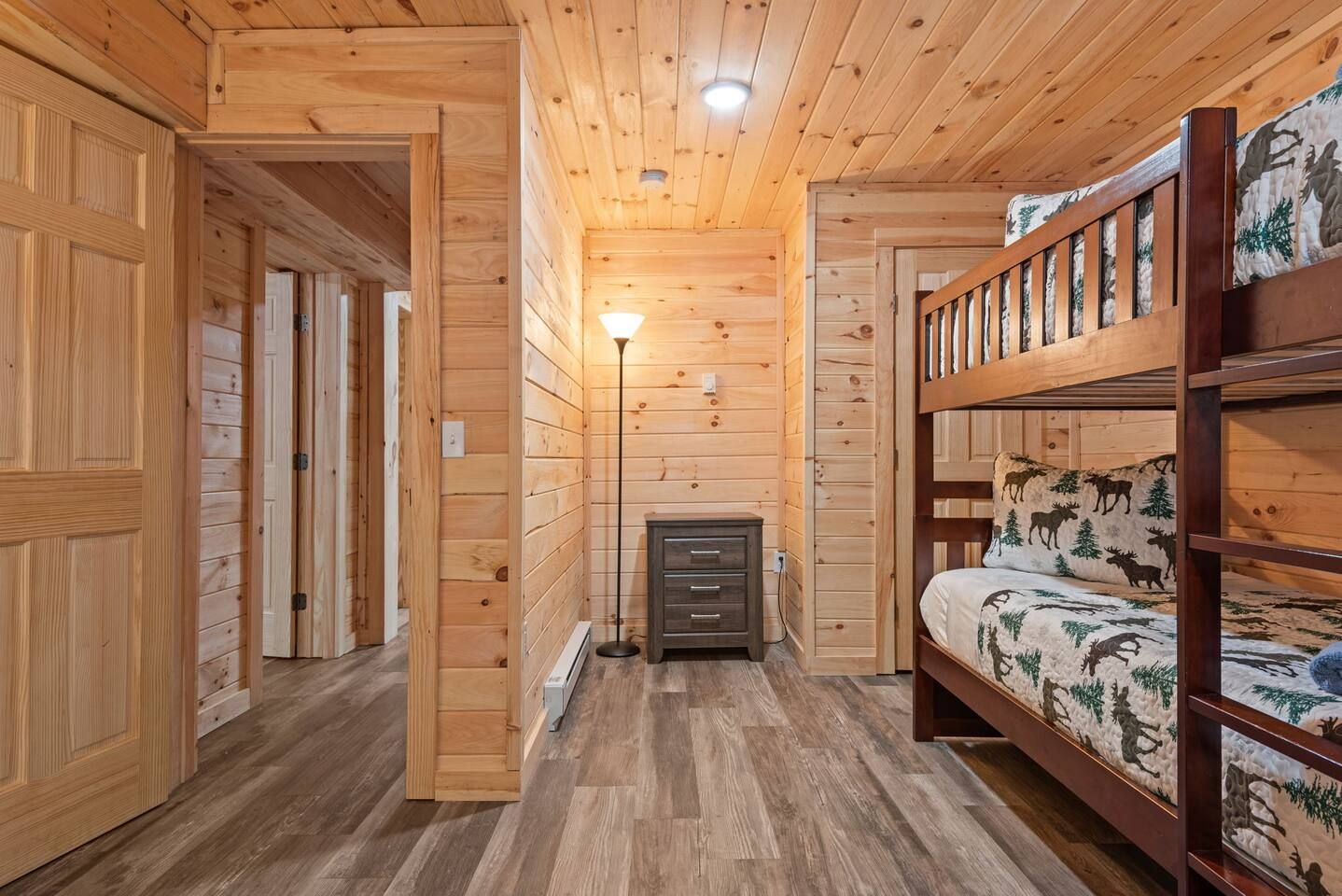 Interior de una cabaña de madera con una cama nido, un aparador y una lámpara de pie.
