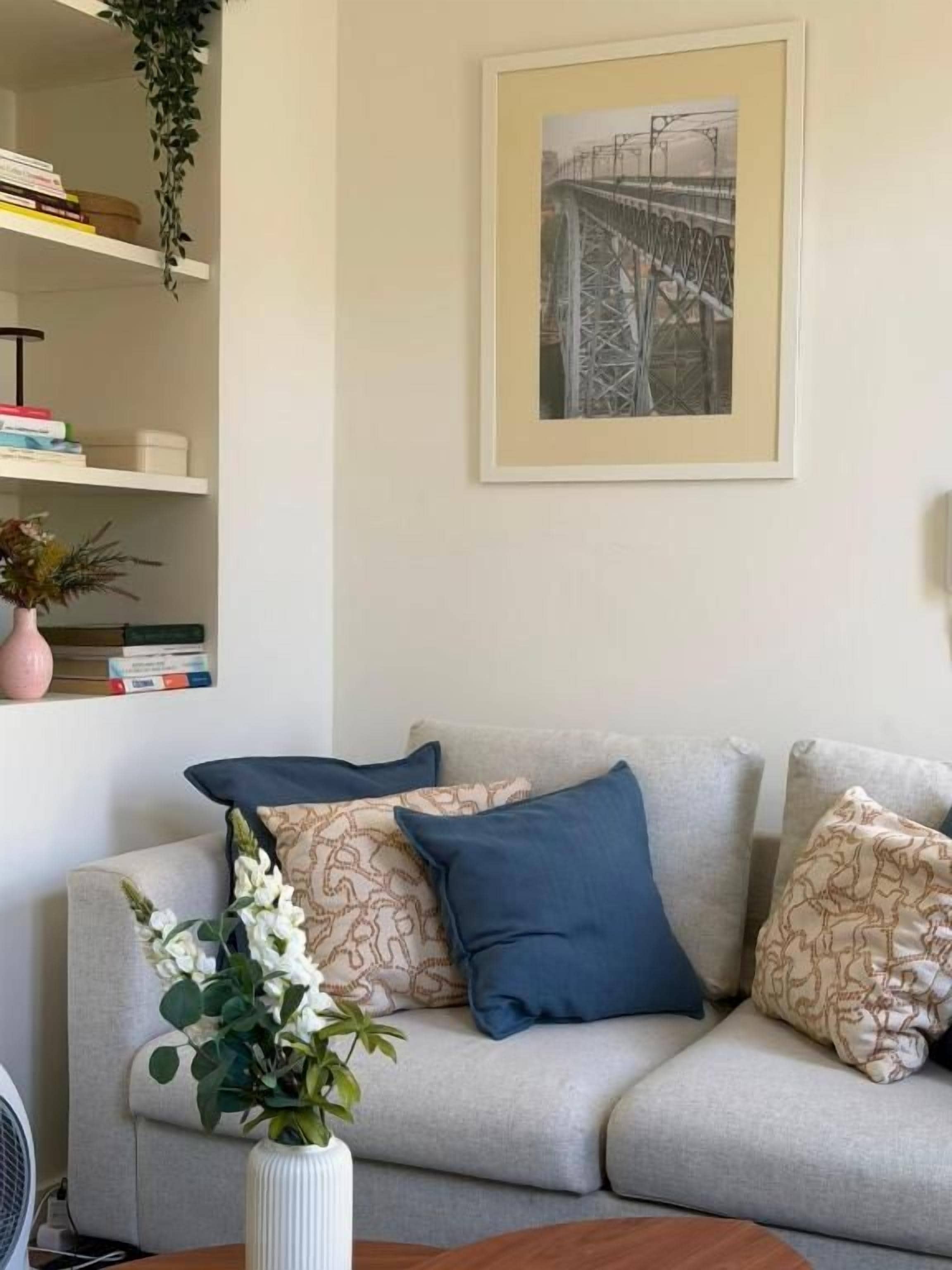 Ein Wohnzimmer mit einer Couch, Kissen, einem gerahmten Bild an der Wand und einem Bücherregal mit Büchern und einer Vase.