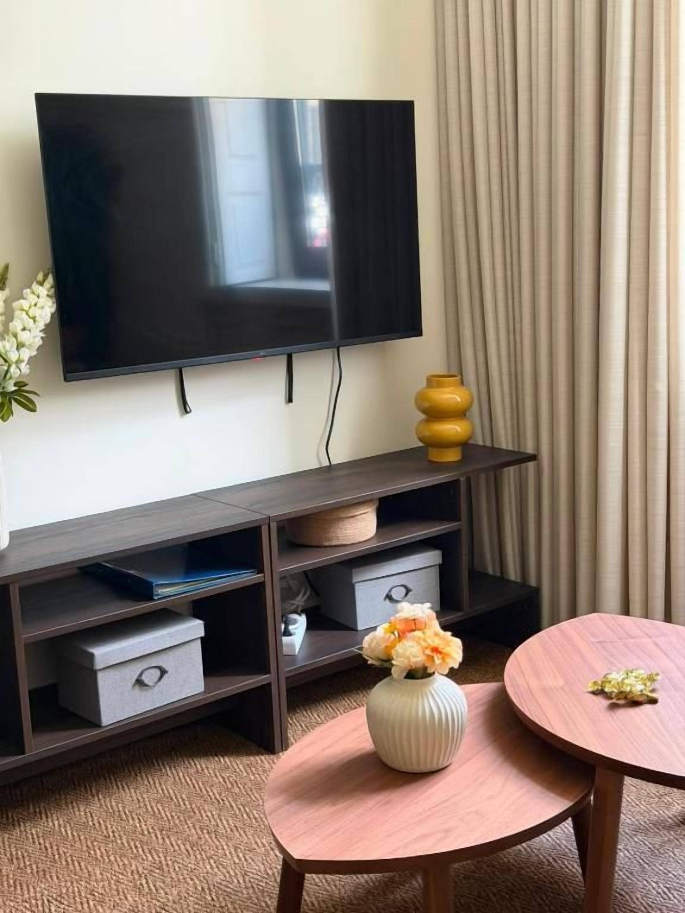 Une pièce dotée d’un téléviseur à écran plat fixé au mur, d’un meuble de rangement en bois servant de support pour les appareils multimédias, d’une table ronde en bois ornée d’un vase de fleurs et d’une fenêtre recouverte d’un rideau.